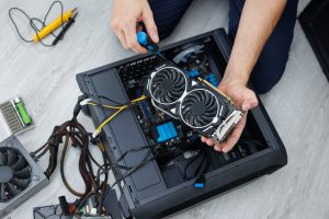 Technician repairing a desktop computer, holding a GPU (graphics card) with tools and cables visible on a workbench.