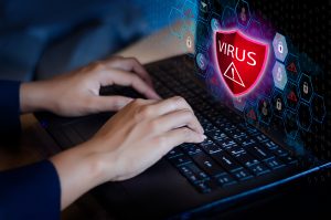Hands typing on a laptop keyboard with a digital shield icon labeled 'Virus' representing cybersecurity and virus protection.