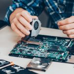 Technician replacing a cooling fan on a laptop motherboard using specialized tools.