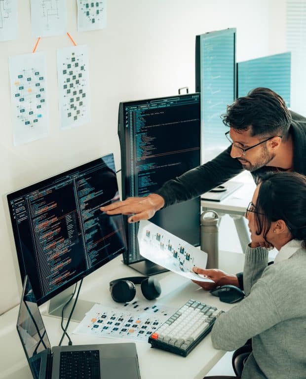 Two IT professionals analyzing software performance and discussing optimization strategies on a multi-monitor setup.